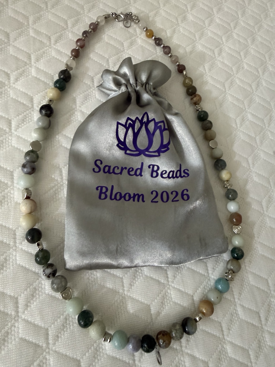 A beaded necklace surrounding a silver Sacred Beads Bloom 2026 keepsake pouch.